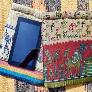 Op de afbeelding: Een tabletcomputer met een zwart scherm bevindt zich in een kleurrijke stoffen hoes met een rood, groen en bruin patroon. De hoes heeft een flap die over de tablet vouwt en is versierd met verschillende kleine, kleurrijke ontwerpen.