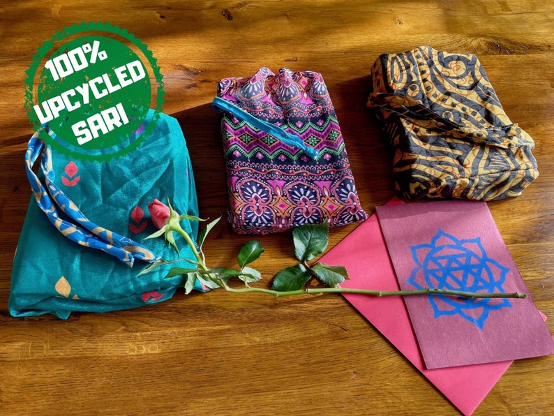 Pu&ograve; includere: Tre pochette in tessuto realizzate con tessuto sari riciclato al 100%. Le pochette sono di colori e motivi diversi. Un'unica rosa rossa &egrave; appoggiata sulle pochette.
