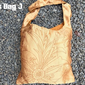 Pu&ograve; includere: Una borsa a tracolla color oro con un motivo floreale. La borsa ha una sola tracolla ed &egrave; realizzata in un materiale setoso. Il testo "Cross Bag J" si trova nell'angolo in alto a sinistra. La borsa &egrave; appoggiata su un letto di ghiaia grigia.