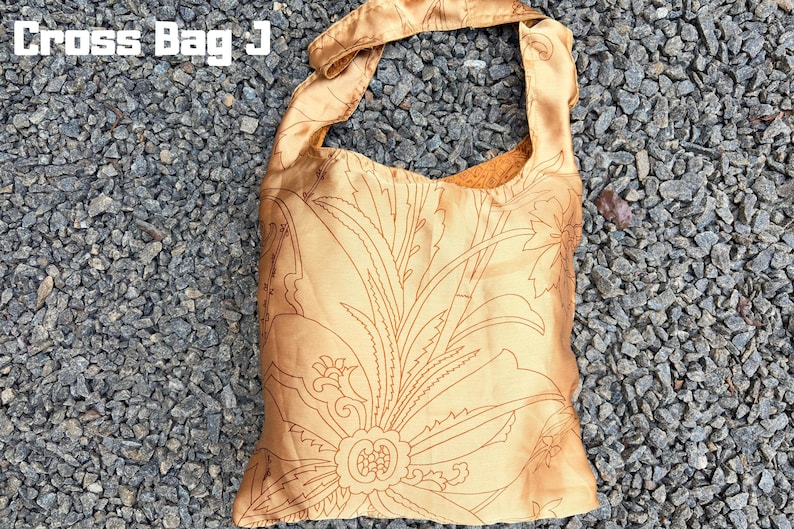 Pu&ograve; includere: Una borsa a tracolla color oro con un motivo floreale. La borsa ha una sola tracolla ed &egrave; realizzata in un materiale setoso. Il testo "Cross Bag J" si trova nell'angolo in alto a sinistra. La borsa &egrave; appoggiata su un letto di ghiaia grigia.