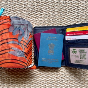 Pu&ograve; includere: Un portapassaporto blu con un passaporto blu all'interno, un passaporto bordeaux e tre carte di credito. Il portapassaporto ha una chiusura a zip e una piccola finestra con il testo "FOLLOW THE FROG" e un codice QR.