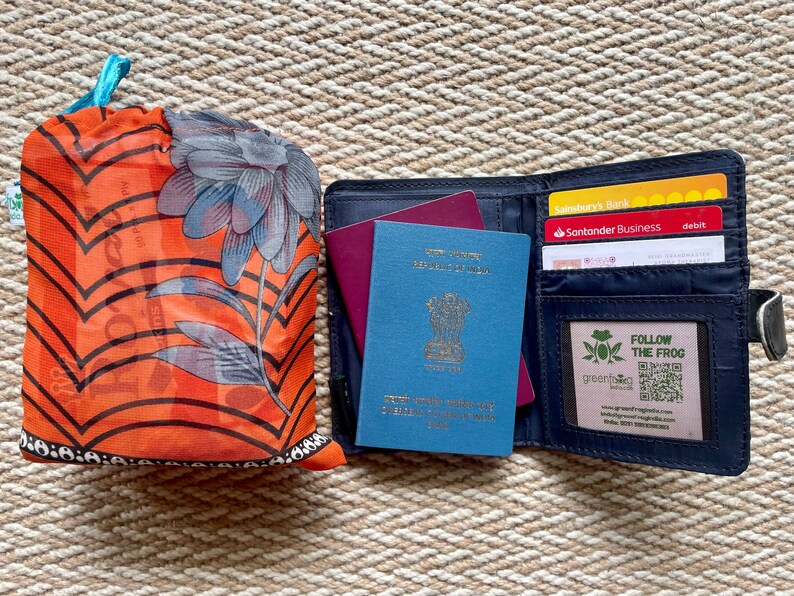 Pu&ograve; includere: Un portapassaporto blu con un passaporto blu all'interno, un passaporto bordeaux e tre carte di credito. Il portapassaporto ha una chiusura a zip e una piccola finestra con il testo "FOLLOW THE FROG" e un codice QR.
