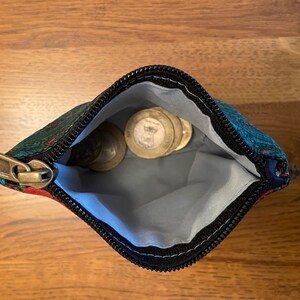 K&ouml;nnte beinhalten: Eine kleine, mit einem Rei&szlig;verschluss versehene Tasche mit einem schwarzen Rei&szlig;verschluss und einem bunten Blumenmuster. Die Tasche ist offen und enth&auml;lt mehrere M&uuml;nzen.