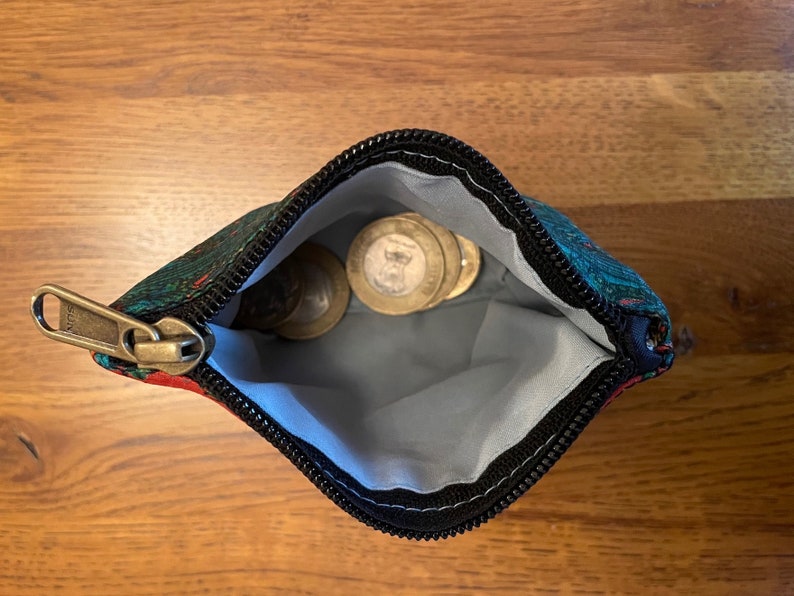 K&ouml;nnte beinhalten: Eine kleine, mit einem Rei&szlig;verschluss versehene Tasche mit einem schwarzen Rei&szlig;verschluss und einem bunten Blumenmuster. Die Tasche ist offen und enth&auml;lt mehrere M&uuml;nzen.
