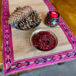 May include: A burlap table runner with a pink and purple ikat border. The runner is decorated with a bowl of red berries and a bowl of pine cones.