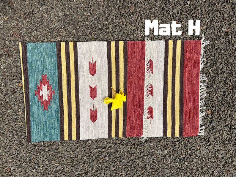 Pu&ograve; includere: Un tappeto a strisce colorate con un motivo geometrico al centro. Il tappeto ha frange sui bordi ed &egrave; fatto di tessuto tessuto. Il tappeto &egrave; etichettato "Mat H".