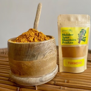 Pode incluir: Uma tigela de madeira cheia de pó de açafrão amarelo brilhante e uma colher de madeira. Uma sacola de papel kraft com um rótulo amarelo que diz "Malabar Spice Mountains Produce" e "Turmeric" está ao lado da tigela. O fundo é um tapete tecido marrom claro.