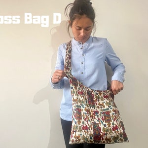 Pu&ograve; includere: Una borsa a tracolla color crema con un motivo colorato di figure e forme. La borsa ha una lunga tracolla ed &egrave; indossata da una persona che indossa una camicia azzurra. Il testo "Cross Bag D" &egrave; visibile nell'immagine.