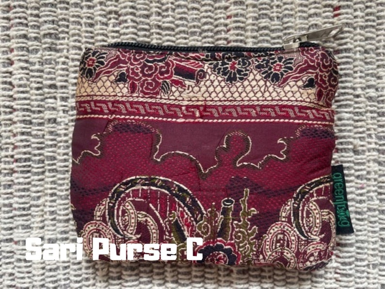 K&ouml;nnte beinhalten: Eine rote Stofftasche mit goldenen Mustern und einem Rei&szlig;verschluss. Die Tasche hat ein gr&uuml;nes Etikett mit dem Wort "greenswag" darauf. Die Tasche ist mit "Sari Purse C" beschriftet.