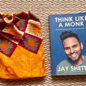 Pu&ograve; includere: Un libro intitolato "Think Like A Monk: Train Your Mind for Peace and Purpose Every Day" di Jay Shetty. La copertina del libro presenta una foto dell'autore, Jay Shetty, sorridente.