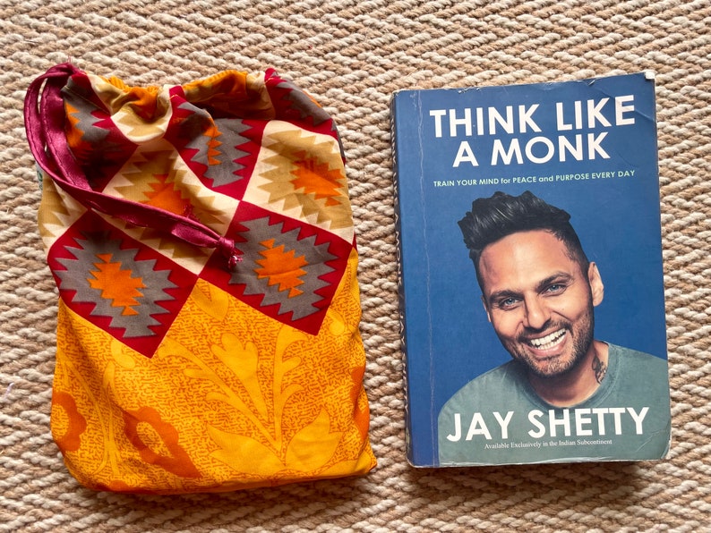 Pu&ograve; includere: Un libro intitolato "Think Like A Monk: Train Your Mind for Peace and Purpose Every Day" di Jay Shetty. La copertina del libro presenta una foto dell'autore, Jay Shetty, sorridente.