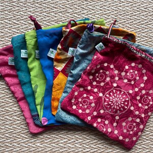 Pu&ograve; includere: Un set di otto sacchetti per prodotti riutilizzabili in tessuto colorati. I sacchetti hanno chiusure con coulisse e presentano diversi motivi e design. I sacchetti sono piegati e disposti a ventaglio su una superficie testurizzata.