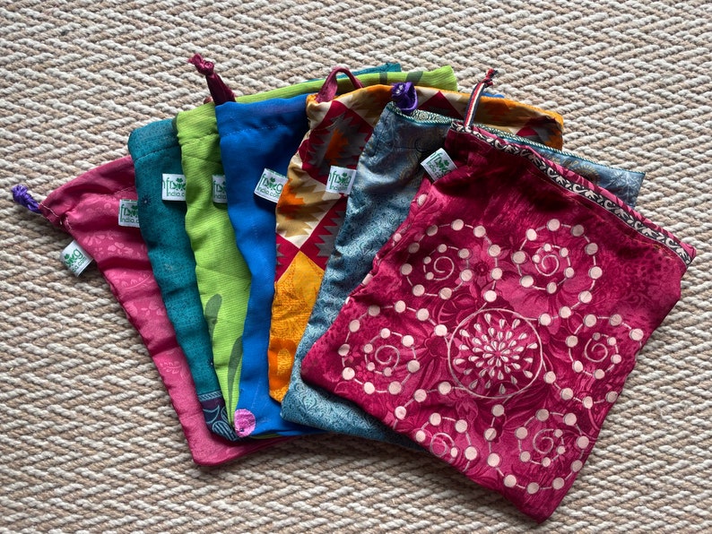 Pu&ograve; includere: Un set di otto sacchetti per prodotti riutilizzabili in tessuto colorati. I sacchetti hanno chiusure con coulisse e presentano diversi motivi e design. I sacchetti sono piegati e disposti a ventaglio su una superficie testurizzata.