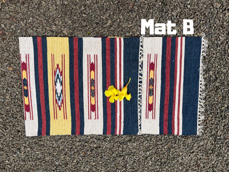Pu&ograve; includere: Un tappeto tessuto con un motivo geometrico in tonalit&agrave; di giallo, rosso, bianco e blu. Il tappeto ha un bordo con frange ed &egrave; etichettato "Mat B".