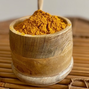 Puede incluir: Un cuenco de madera lleno de polvo de cúrcuma amarillo brillante. El cuenco tiene una veta de madera natural y un color marrón claro. La cúrcuma está finamente molida y tiene un tono dorado vibrante. El cuenco está sobre una superficie de bambú.
