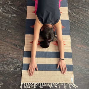 Pu&ograve; includere: Una persona che esegue una posa di yoga su un tappeto a strisce con strisce blu e beige. Il tappeto ha un bordo a frangia.