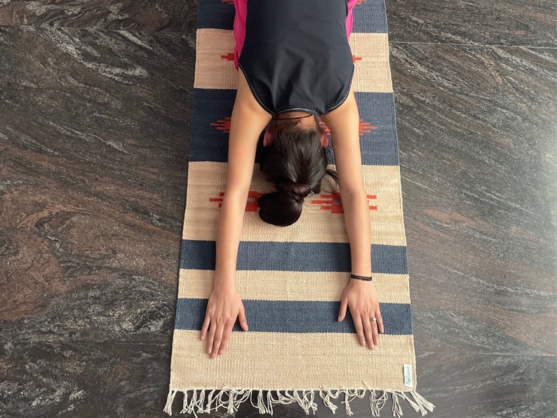 Pu&ograve; includere: Una persona che esegue una posa di yoga su un tappeto a strisce con strisce blu e beige. Il tappeto ha un bordo a frangia.