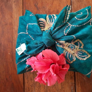 Puede incluir: Un envoltorio de tela turquesa con detalles de lentejuelas doradas y una flor de hibisco rosa. La tela está anudada y presenta motivos florales. El envoltorio tiene una etiqueta con el texto "mkgdia.com".