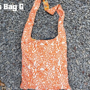 Pu&ograve; includere: Borsa a tracolla arancione e bianca con un motivo geometrico ripetuto. La borsa ha una lunga tracolla e il testo "Cross Bag G" in lettere bianche in alto. La borsa &egrave; in tessuto e poggia su un letto di piccole pietre grigie.
