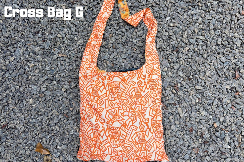 Pu&ograve; includere: Borsa a tracolla arancione e bianca con un motivo geometrico ripetuto. La borsa ha una lunga tracolla e il testo "Cross Bag G" in lettere bianche in alto. La borsa &egrave; in tessuto e poggia su un letto di piccole pietre grigie.