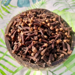 Può includere: Una ciotola di legno piena di chiodi di garofano interi. I chiodi di garofano sono di colore marrone scuro e hanno una piccola testa arrotondata. La ciotola è posta su uno sfondo con un motivo floreale bianco e verde.
