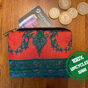 K&ouml;nnte beinhalten: Eine kleine, rot-blaue, gemusterte Tasche mit Rei&szlig;verschluss. Die Tasche ist aus 100 % recyceltem Sari-Stoff gefertigt. Die Tasche liegt auf einer Holzoberfl&auml;che mit einer Kreditkarte und M&uuml;nzen darin.