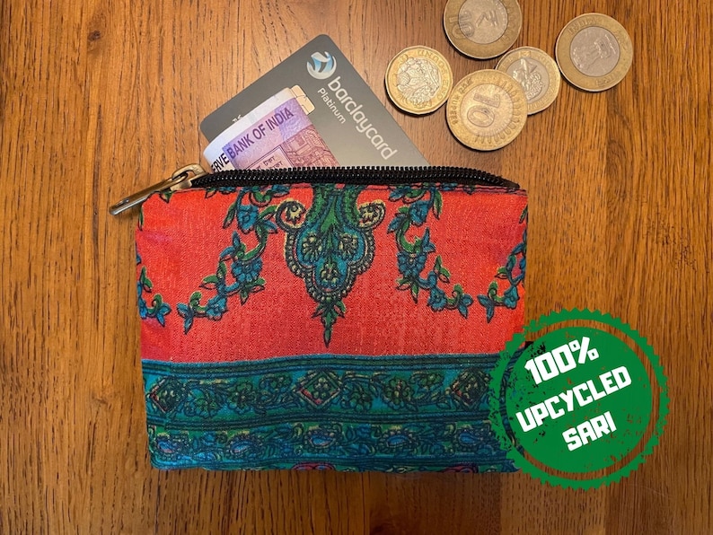 K&ouml;nnte beinhalten: Eine kleine, rot-blaue, gemusterte Tasche mit Rei&szlig;verschluss. Die Tasche ist aus 100 % recyceltem Sari-Stoff gefertigt. Die Tasche liegt auf einer Holzoberfl&auml;che mit einer Kreditkarte und M&uuml;nzen darin.