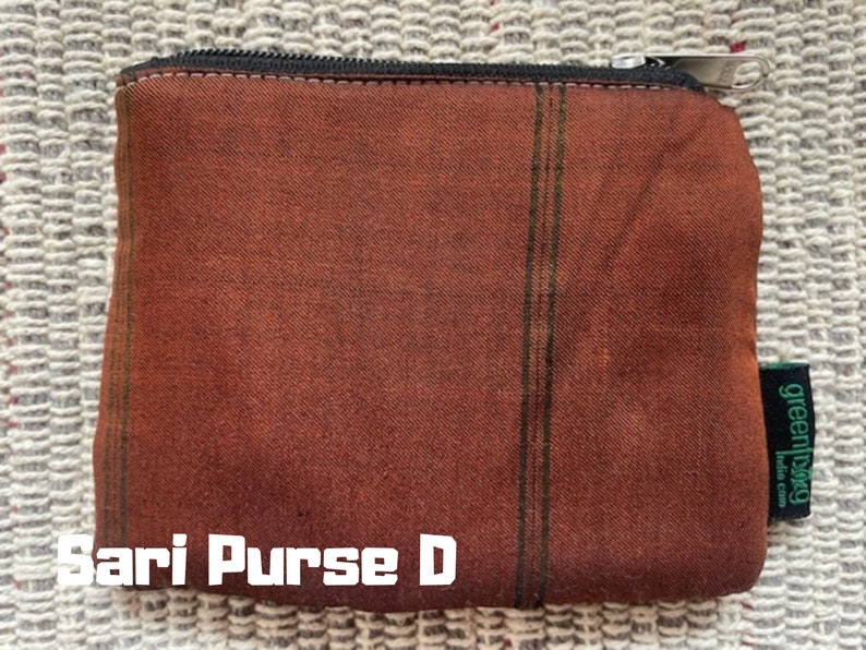 K&ouml;nnte beinhalten: Eine braune und gr&uuml;n gestreifte Stofftasche mit Rei&szlig;verschluss. Die Tasche hat ein gr&uuml;nes Etikett mit der Aufschrift "greenbag India Craft". Der Text "Sari Purse D" ist auf der Vorderseite der Tasche aufgedruckt.