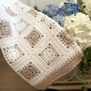 May include: A crocheted baby blanket with a white and beige pattern. The blanket is made of soft yarn and has a delicate, lacy design.
