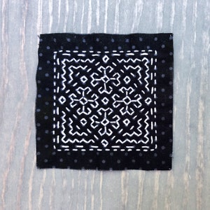 May include: Square black fabric with white embroidered geometric design. The fabric has a border of small white polka dots. The central design is a complex pattern of white lines and shapes.