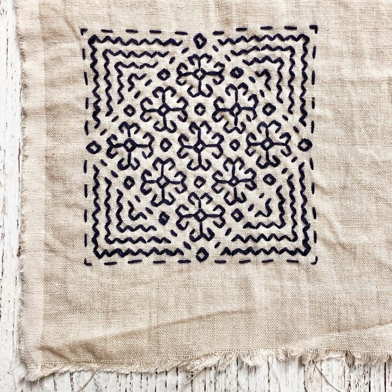 May include: A square of beige fabric with a dark blue embroidered design. The design features a geometric pattern within a square border, with a raw, unfinished edge. The embroidery is detailed and intricate.