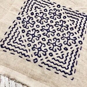 May include: Close-up of a beige fabric with a dark blue embroidered design. The design is a square pattern with floral and geometric shapes. The edges of the fabric are unfinished, with visible stitching.