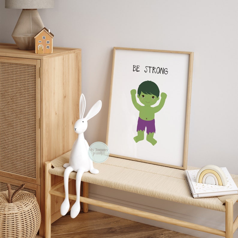 Op de afbeelding: Een printbaar wanddecor ontwerp met een groen cartoonfiguur met de tekst "BE STRONG". Het figuur draagt een paarse broek en heeft zijn armen in de lucht gestoken.