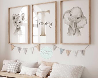 Könnte beinhalten: Drei gerahmte Aquarelldrucke mit einem Löwenjungen, dem Buchstaben „T“ mit dem Namen „Tommy“ und einem Elefanten. Die Drucke sind in neutralen Tönen gehalten und werden über einer Bank mit Kissen und Aufbewahrungsboxen ausgestellt. Unter den Rahmen hängt eine Girlande aus kleinen Flaggen.