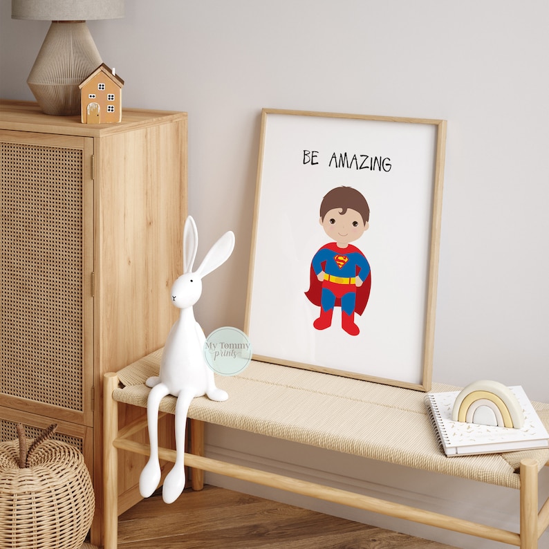 Op de afbeelding: Een ingelijst print met een witte achtergrond en een cartoonillustratie van een superheldenjongen in een rood-blauw kostuum. De tekst "BE AMAZING" is in het zwart bovenaan de print gedrukt.