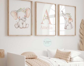 May include: Three framed wall art prints featuring watercolor illustrations of baby elephants with floral crowns. The center print displays a large letter A with a floral accent and the name Amelia printed in a soft pink serif font.