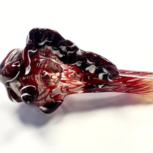 May include: A glass pipe shaped like a seashell with red, white, and black swirling patterns.