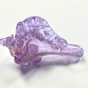 May include: A handcrafted, abstract glass sculpture in shades of lavender and white. The art glass piece has a unique, organic shape with a smooth, glossy finish. The sculpture is approximately 8 inches long.