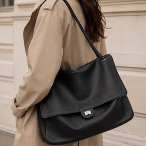 Puede incluir: Un bolso de hombro grande de color negro con textura granulada, que presenta una solapa, un cierre giratorio rectangular de tono plateado y una correa de hombro única sujeta con herrajes plateados, llevado sobre una gabardina beige.