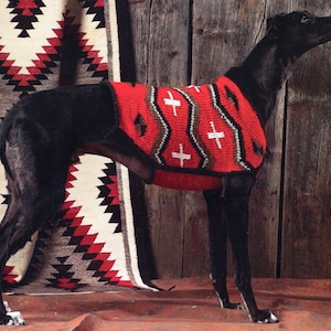 May include: A black Greyhound dog wearing a red, brown, and white knitted sweater with a geometric pattern. The dog is standing on a red and white patterned rug.