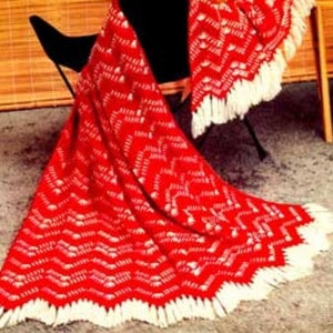 May include: A red and white crocheted blanket with a chevron pattern and white fringe. The blanket is draped over a black chair. The blanket is a cozy home decor item.