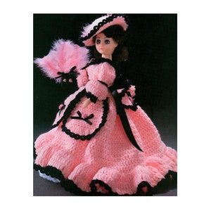 May include: A vintage doll in a pink crochet dress with black trim, a matching hat, and a pink feather fan. The doll has blue eyes and dark hair. The dress features a ruffled skirt and black ribbon accents, creating a classic, handmade aesthetic.