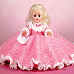 May include: A doll with blonde hair and blue eyes wearing a pink crocheted dress. The dress features a wide, flared skirt with a white ruffled trim at the hem, white ruffled cuffs on the sleeves, and a white ruffled collar with a small pink bow.