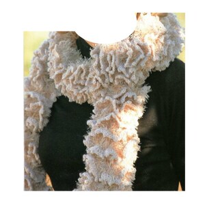 May include: A beige crocheted scarf with a ruffled texture. The scarf is worn around the neck of a person wearing a black shirt.