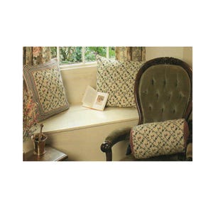 May include: An interior shot featuring a cozy reading nook. A green velvet armchair is next to a window with floral curtains. Decorative pillows with a floral pattern and a bolster pillow are on the window seat and chair. A small book is open.