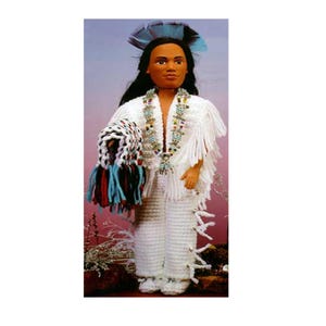 May include: A handmade doll dressed in a Native American-inspired outfit. The doll wears a white crocheted outfit with fringe, beaded necklaces, and a feathered headdress. The doll is holding a fringed bag with blue, red, and black accents.