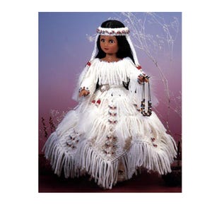 May include: A handcrafted doll wearing a white, fringed dress with red and blue accents. The doll has dark hair and a beaded headband. It holds a beaded necklace. The background is a gradient of purple and pink.