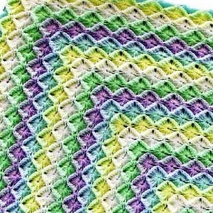 May include: A handmade, square crochet blanket in a vibrant array of colours. The blanket features a repeating pattern of textured stitches in shades of green, yellow, purple, blue, and white. The edges are finished with a turquoise border.