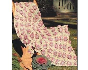May include: A crocheted throw blanket with a floral pattern in shades of pink and white. The blanket is draped over a wooden chair and is surrounded by green grass.