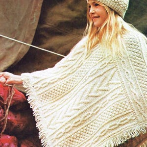 May include: A white knitted poncho with a fringe border and a matching white knitted hat with a pom pom on top.